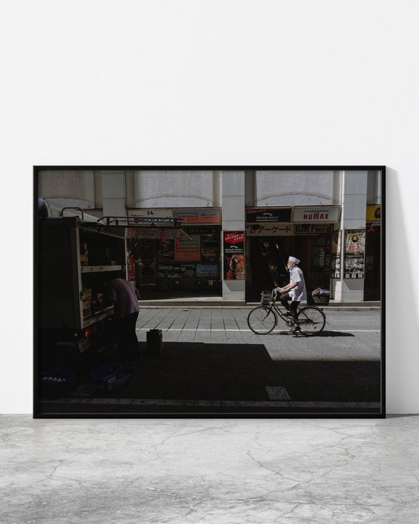 Urban street scene with a cyclist passing storefronts, contrasted by deep shadows and modern city textures.