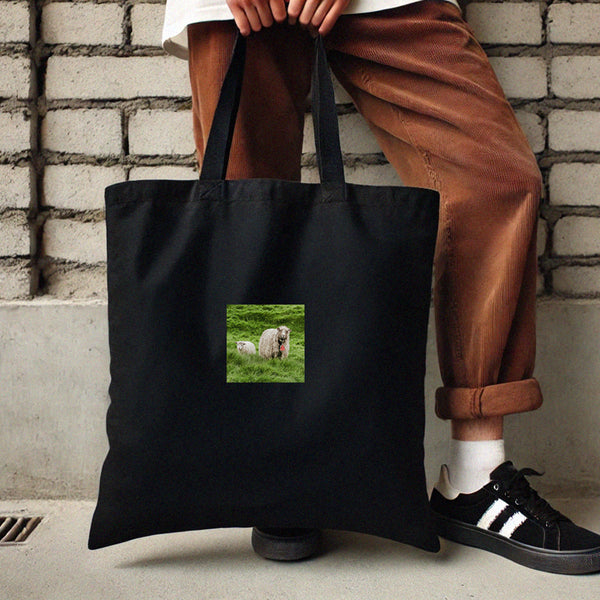 Model holding a black tote bag with a centered sheep and lamb print, styled with casual streetwear.