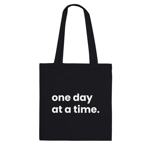 A black canvas tote bag designed by North Minimal featuring white letter text that reads "one day at a time.". The image shows the front of the tote bag with the text centered and clearly visible on the black background.