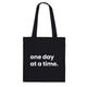 A black canvas tote bag designed by North Minimal featuring white letter text that reads "one day at a time.". The image shows the front of the tote bag with the text centered and clearly visible on the black background.